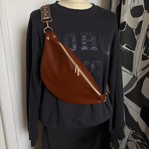 Könnte beinhalten: Eine braune Bauchtasche mit goldenem Reißverschluss und Beschlägen wird auf einer Schaufensterpuppe präsentiert. Die Tasche hat einen gemusterten Riemen und wird mit einem schwarzen Sweatshirt und Rock kombiniert. Auf dem Sweatshirt steht das Wort "SOHO".