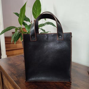 Può includere: Una borsa tote in pelle nera con cuciture e manici marroni. La borsa ha una forma quadrata ed è appoggiata su una superficie di legno. I manici sono fissati con rivetti. La borsa è vuota.