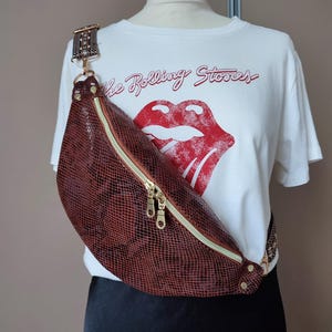 May include: A brown snakeskin-effect bum bag with a gold zip and hardware is displayed on a mannequin. The bag is worn across the body. A white t-shirt with "The Rolling Stones" and a red tongue logo is visible.
