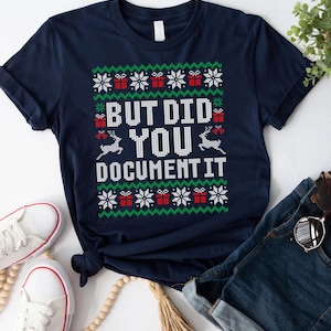 May include: A navy blue t-shirt with a Christmas-themed design. The shirt displays the text "BUT DID YOU DOCUMENT IT" in white, with festive snowflake, gift box, and reindeer motifs. The shirt is shown with white trainers and denim jeans.