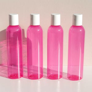 May include: Four clear, pink plastic bottles with white flip-top caps are arranged in a row. The bottles are cylindrical and appear to be empty. The background is a light pink color.