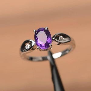 May include: A silver-toned ring featuring a central oval-cut purple amethyst gemstone held in a prong setting. The band includes two smaller, clear round-cut accent stones, one on each side of the central amethyst, set within the metal band.