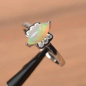 May include: A silver ring featuring a marquise-cut opal gemstone with a pearlescent sheen. The opal is surrounded by small, clear accent stones set in a floral design. The ring's band is a polished silver.
