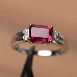 Pode incluir: Um anel de prata com uma pedra preciosa retangular, vermelho escuro, como peça central. O anel é adornado com pedras preciosas menores e transparentes em ambos os lados da pedra principal, criando um design clássico e elegante. A banda tem um acabamento liso e polido.