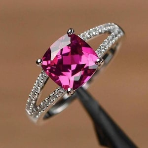 Può includere: Un anello d'argento con una grande pietra preziosa rosa brillante di forma quadrata come elemento centrale. La fascia si divide in due, ciascuna con piccole pietre trasparenti. L'anello è ambientato su uno sfondo sfocato.