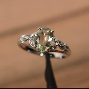 May include: A silver ring with a pale green oval gemstone. The ring has a decorative band with leaf-shaped patterns. The central gemstone is the main feature, set in a prominent position.