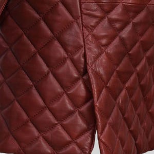 K&ouml;nnte beinhalten: Nahaufnahme einer burgunderfarbenen, gesteppten Lederjacke. Die Jacke hat ein Rautenmuster und einen Schlitz am unteren Saum. Das Leder hat einen leichten Glanz, und die N&auml;hte sind sichtbar.