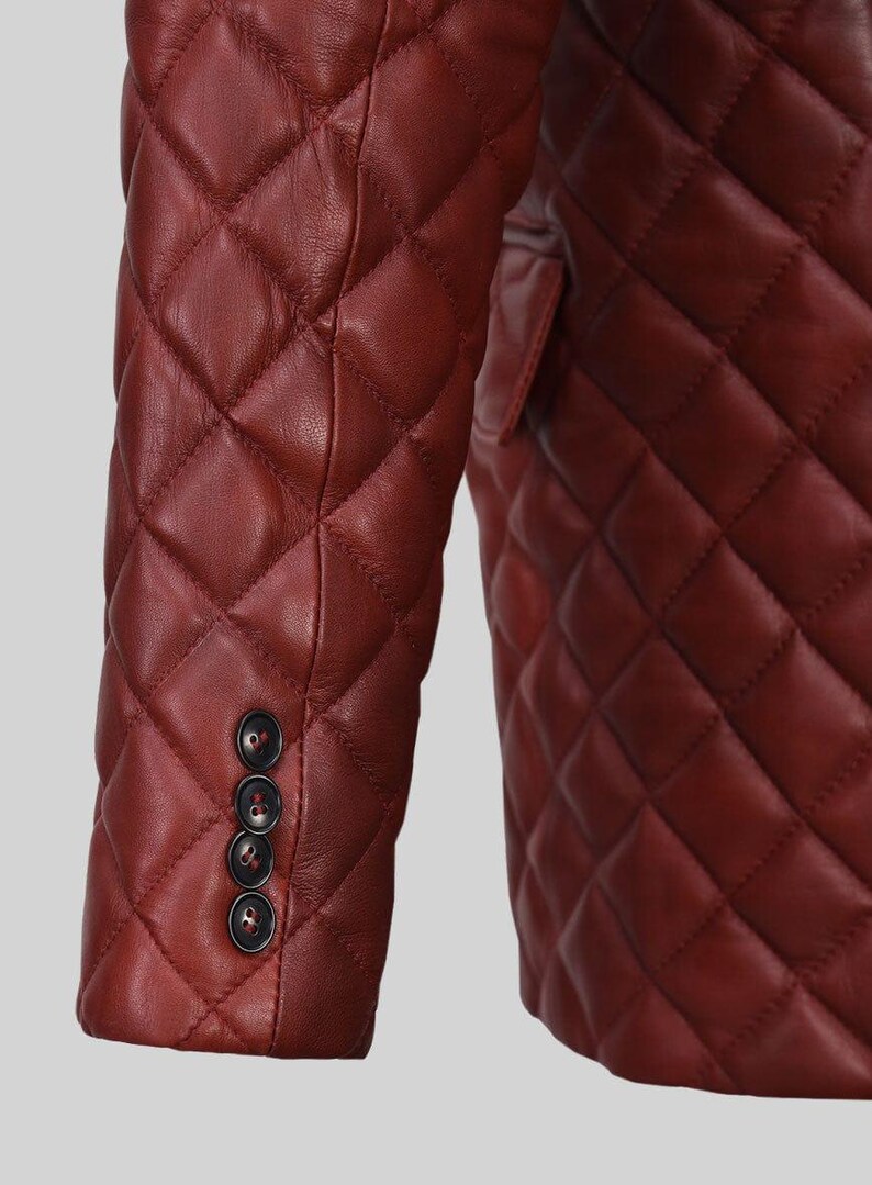 K&ouml;nnte beinhalten: Nahaufnahme einer burgunderfarbenen, gesteppten Lederjacke. Die Jacke hat ein Rautenmuster und drei dunkle Kn&ouml;pfe am &Auml;rmel. Das Design der Jacke deutet auf einen klassischen, ma&szlig;geschneiderten Stil hin.