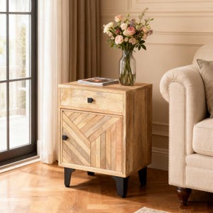 May include: A light-colored wooden nightstand with a drawer and a door featuring a geometric design. The nightstand has black hardware and legs. A glass vase with flowers sits on top, and a magazine rests on the nightstand.