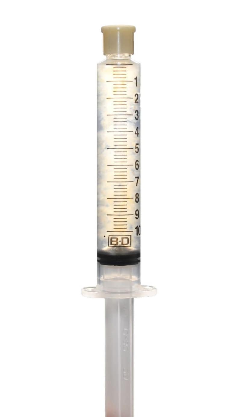 May include: A clear plastic syringe with a beige cap and a black and white label that reads "B-D". The syringe is filled with a clear liquid and has markings up to 10 milliliters.