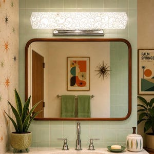 May include: A rectangular wall mirror with a rounded wooden frame, positioned above a bathroom sink. The mirror reflects a wall with a framed abstract print, a towel rack with two green towels, and a metal starburst wall decoration.