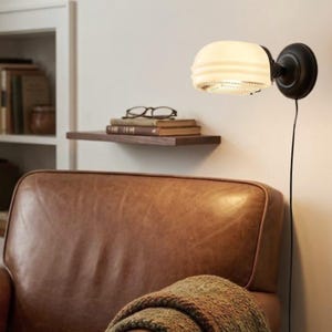 May include: A wall-mounted lamp with a cream-colored, ribbed glass shade and a black metal fixture. The lamp is positioned above a brown leather armchair, with a small wooden shelf holding books and eyeglasses in the background.