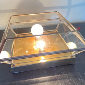 May include: A vintage-style table lamp with a geometric glass and brass frame. The lamp features a square design with clear glass panels and a brass base. Three light bulbs are visible, illuminating the interior. The lamp is plugged in and ready to use.