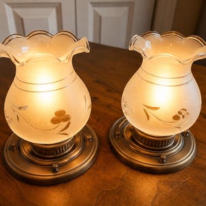 May include: A pair of matching wall sconces featuring frosted glass shades with a scalloped rim and etched floral patterns. The shades are illuminated from within and are mounted on circular, metallic bronze-toned bases with a ribbed design.