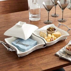 May include: A white ceramic double serving tray with a metal frame and handles. One side holds a stack of light blue napkins with silver picks, while the other side contains cubes of cheese and a small bowl of olives. A white lid sits partially open.