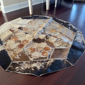 May include: An octagonal patchwork rug with a cowhide design. The rug features various shades of brown, tan, and black, with visible stitching. The rug is placed on a dark wood floor.