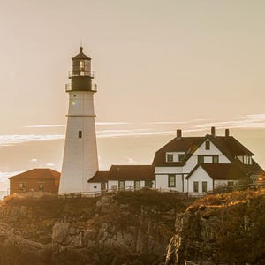 Puede incluir: Un faro blanco con una parte superior negra se alza sobre un acantilado rocoso, junto a un edificio blanco con tejado rojo. La escena está bañada por la cálida luz del sol, con el mar visible a lo lejos.