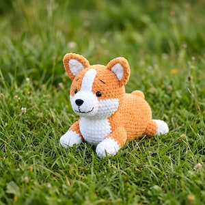 Pode incluir: Um brinquedo de pel&uacute;cia de cachorro corgi de croch&ecirc; em laranja, branco e preto, deitado na grama verde. O animal de pel&uacute;cia feito &agrave; m&atilde;o tem um rosto sorridente, orelhas ca&iacute;das e patas brancas. O brinquedo &eacute; um item macio e fofinho.