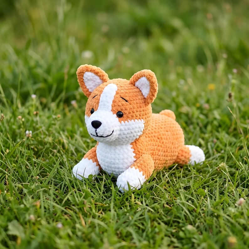 Pode incluir: Um brinquedo de pel&uacute;cia de cachorro corgi de croch&ecirc; em laranja, branco e preto, deitado na grama verde. O animal de pel&uacute;cia feito &agrave; m&atilde;o tem um rosto sorridente, orelhas ca&iacute;das e patas brancas. O brinquedo &eacute; um item macio e fofinho.