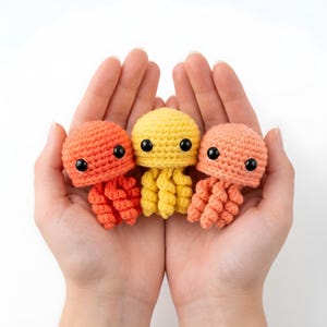 Pode incluir: Tr&ecirc;s &aacute;guas-vivas de croch&ecirc; feitas &agrave; m&atilde;o em tons de laranja, amarelo e p&ecirc;ssego. Cada &aacute;gua-viva tem uma cabe&ccedil;a redonda com olhos pretos grandes e tent&aacute;culos encaracolados. As &aacute;guas-vivas s&atilde;o seguradas em m&atilde;os em concha sobre um fundo branco.