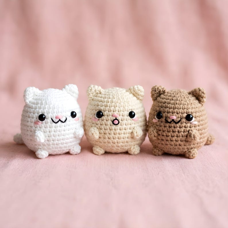 Pode incluir: Tr&ecirc;s brinquedos de gato de croch&ecirc; em branco, bege e marrom. Cada um tem um corpo redondo, orelhas pequenas e um rosto fofo com olhos pretos e bochechas rosadas. Os brinquedos est&atilde;o dispostos em um fundo rosa.