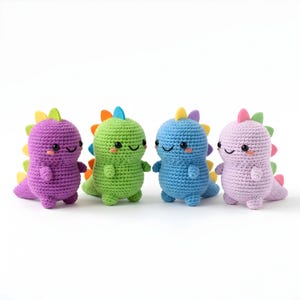 Pode incluir: Quatro brinquedos de dinossauro de croch&ecirc; em roxo, verde, azul e rosa. Cada dinossauro tem um rosto sorridente, bra&ccedil;os pequenos e uma espinha colorida. Os brinquedos s&atilde;o feitos &agrave; m&atilde;o e ficam em p&eacute;.