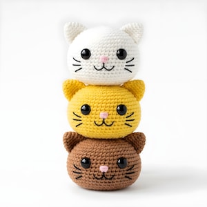 Pode incluir: Tr&ecirc;s cabe&ccedil;as de gato de croch&ecirc; empilhadas em branco, amarelo e marrom. Cada cabe&ccedil;a de gato tem olhos pretos, narizes rosa e bigodes bordados. Os brinquedos de gato feitos &agrave; m&atilde;o s&atilde;o empilhados.