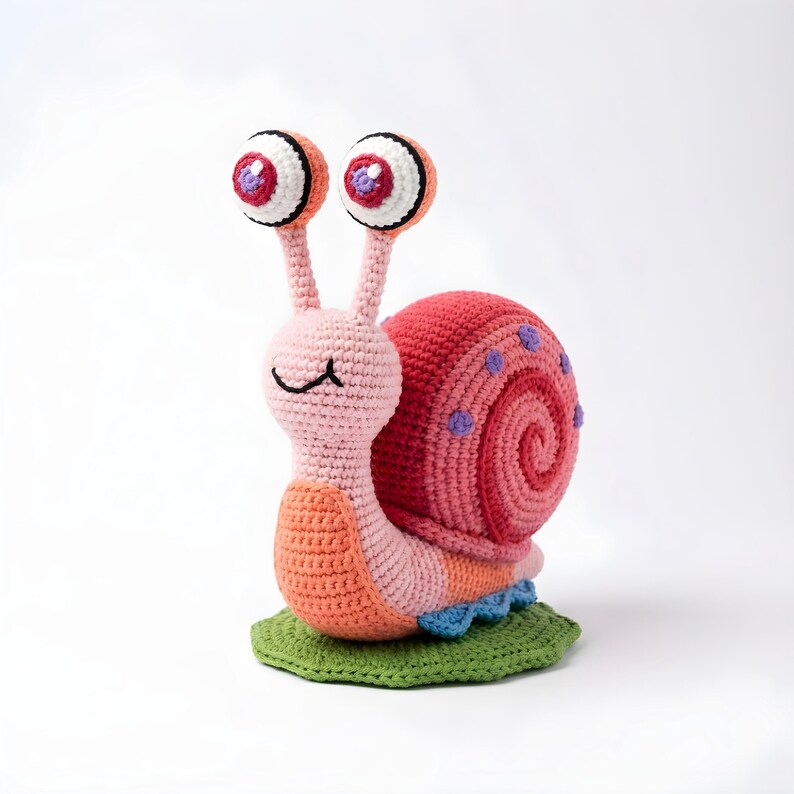 Pode incluir: Um brinquedo de caracol de croch&ecirc; feito &agrave; m&atilde;o com olhos grandes e expressivos. O caracol apresenta um corpo rosa, uma concha vermelha com manchas roxas e senta-se em uma folha verde. O design do caracol &eacute; l&uacute;dico e colorido, perfeito para um quarto infantil.