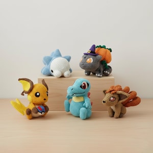 Puede incluir: Colección de cinco figuras de Pokémon de ganchillo en varios colores. Las figuras incluyen una criatura amarilla y marrón sosteniendo una pelota, un dinosaurio azul, una criatura parecida a un zorro marrón, una criatura azul y blanca y una criatura gris con un sombrero de bruja.
