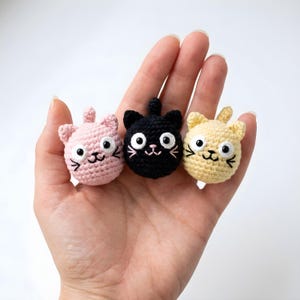 Pode incluir: Tr&ecirc;s cabe&ccedil;as de gato de croch&ecirc; em rosa, preto e amarelo. Cada gato tem olhos brancos grandes, um nariz bordado rosa e bigodes bordados pretos. Os gatos t&ecirc;m orelhas pequenas e arredondadas e uma pequena cauda no topo da cabe&ccedil;a.