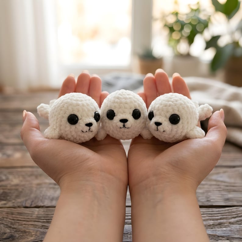 Pode incluir: Tr&ecirc;s focas brancas de croch&ecirc; feitas &agrave; m&atilde;o com olhos pretos e pequenos sorrisos pretos s&atilde;o seguradas em m&atilde;os abertas. As focas t&ecirc;m pequenos ap&ecirc;ndices semelhantes a barbatanas. O fundo est&aacute; desfocado, com luz natural e vegeta&ccedil;&atilde;o vis&iacute;vel.
