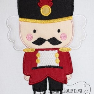 May include: A red and white embroidered nutcracker with a black hat and a gold trim. The nutcracker has a white beard, black mustache, and pink cheeks.
