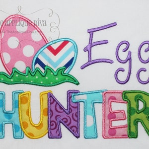 Puede incluir: Diseño bordado con dos huevos de Pascua, uno rosa con lunares blancos y el otro azul y blanco a rayas, sentados sobre hierba verde. El texto "Egg Hunter" está bordado en letras de colores.