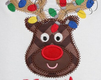 May include: Embroidered reindeer design with antlers adorned with colorful Christmas lights. The reindeer has a red nose, brown and white eyes, and the name "Maggie" in red script below. The Applique Diva logo is in the top center.