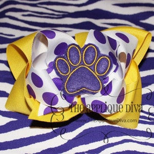 May include: A yellow and white polka dot hair bow with a purple paw print applique. The bow is on a purple and white zebra print background. The text "The Applique Diva" is visible in the bottom right corner of the image.