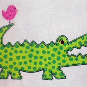 May include: Green alligator with white polka dots and a pink bird on its head. The alligator has a wide open mouth and is made of fabric.