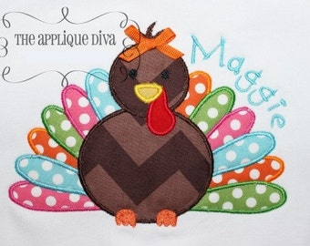 Puede incluir: Un pavo marrón y blanco con una papada roja y una cola de lunares de colores. El pavo tiene un patrón de chevron marrón en su cuerpo y un lazo rojo en su cabeza. El nombre "Maggie" está bordado en azul en el lado derecho de la imagen.