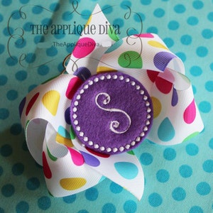 May include: A white ribbon bow with colorful polka dots and a purple felt circle with a white embroidered letter "S" in the center. The bow is on a blue polka dot background.