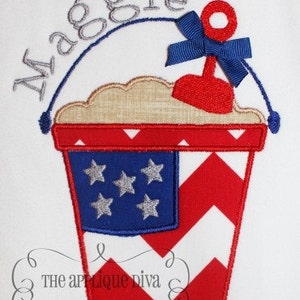 May include: Embroidered design of a red, white and blue bucket with a red shovel and a blue bow. The bucket has a white chevron pattern and a blue section with white stars. The text "Maggie" is embroidered above the bucket.