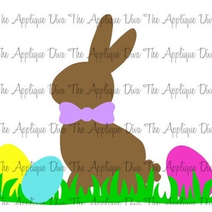 May include: A brown chocolate bunny with a purple bow tie sitting in a patch of green grass with three Easter eggs. The eggs are yellow, blue, and pink.