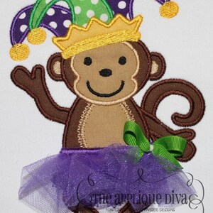 May include: A brown monkey wearing a purple, green, and yellow jester hat with a gold crown. The monkey is wearing a purple tulle tutu and a green bow. The monkey is smiling.