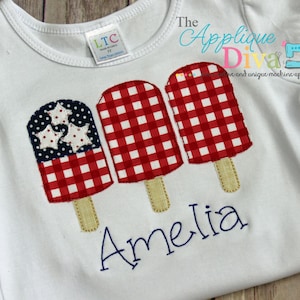May include: A white t-shirt with three red, white, and blue popsicle appliques. The popsicles have a gingham pattern and one has a blue and white star pattern. The name Amelia is embroidered in blue below the popsicles.