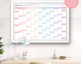 May include: A framed 2026 annual planner with a white border, hanging on a textured wall. The planner is filled with colorful blocks for each month. Text on the planner reads "2026 Annual Planner". A desk with a lamp and office supplies is in front of the planner.