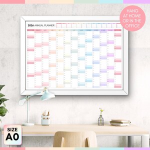 May include: A framed 2026 annual planner with a white border. The planner is filled with colorful blocks for each month and day. Text on the planner reads "2026 Annual Planner." Additional text in a pink circle says "Hang at home or in the office." The size is A0.