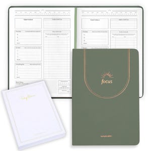 May include: A green planner with the word "focus" in gold lettering and a sunburst design. The planner is open to reveal lined pages for daily scheduling and goal setting. A white notebook with gold lettering is also visible.