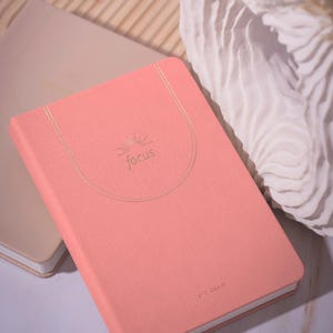 May include: A coral-colored notebook with the word "focus" in gold lettering. The notebook has a gold-colored elastic band and a matching gold-colored sunburst design. The notebook is lying on top of another notebook.