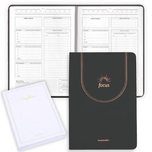 May include: An open, brown, spiral-bound planner with daily and weekly sections, alongside a black notebook with the word "focus" in gold. A white notebook with gold lettering is also present. The planner is designed for organization and productivity.