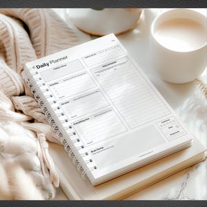 May include: A white spiral-bound daily planner with the words "Daily Planner" at the top. The planner includes sections for priorities, daily schedule, and a brain dump. A mug and a folded sweater are in the background.