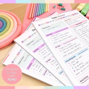 May include: A stack of white daily planner pads with pink and purple accents. Each pad features sections for daily schedules, priorities, and notes. The text "Designed to help with ADHD" is visible on a pink circle.