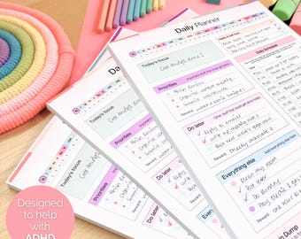 May include: A stack of white daily planner pads with pink and purple accents. Each pad features sections for daily schedules, priorities, and notes. The text "Designed to help with ADHD" is visible on a pink circle.
